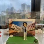 透明壓克力展示盒內，一尊短髮戴眼鏡的人像公仔站在綠色草地質感底座上，穿淺灰外套、深色上衣、白色九分褲與白鞋，右手比讚、左手插口袋。背板是一張校園建築階梯的照片，左上角印有「The Hong Kong Polytechnic University／香港理工大學」及紅色結形校徽，右上角標示「College of Professional and ...」等英文字樣。底座右前角有一小叢多肉植物與石頭裝飾；盒外背景透出城市高樓景色。