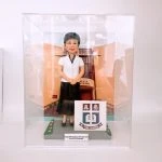 透明壓克力展示盒內，一尊女性人像公仔站在灰色地磚場景中央，穿白色短袖上衣、黑色長裙與黑色平底鞋，短髮、配戴眼鏡並面帶微笑，雙手於身前交握。背板是杏橙色外牆與拱形入口的學校或機構建築。公仔右側有一個立方形立牌，印有校徽與「TSUNG TSIN SCHOOL」字樣；底座前緣的小牌寫着「Happy Retirement to Principal TAM From 2022 Graduates」。地面兩側以綠地與小花點綴。