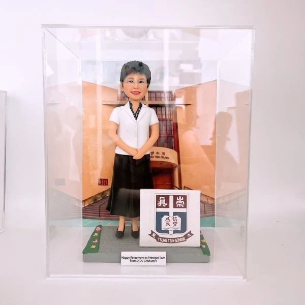 透明壓克力展示盒內，一尊女性人像公仔站在灰色地磚場景中央，穿白色短袖上衣、黑色長裙與黑色平底鞋，短髮、配戴眼鏡並面帶微笑，雙手於身前交握。背板是杏橙色外牆與拱形入口的學校或機構建築。公仔右側有一個立方形立牌，印有校徽與「TSUNG TSIN SCHOOL」字樣；底座前緣的小牌寫着「Happy Retirement to Principal TAM From 2022 Graduates」。地面兩側以綠地與小花點綴。