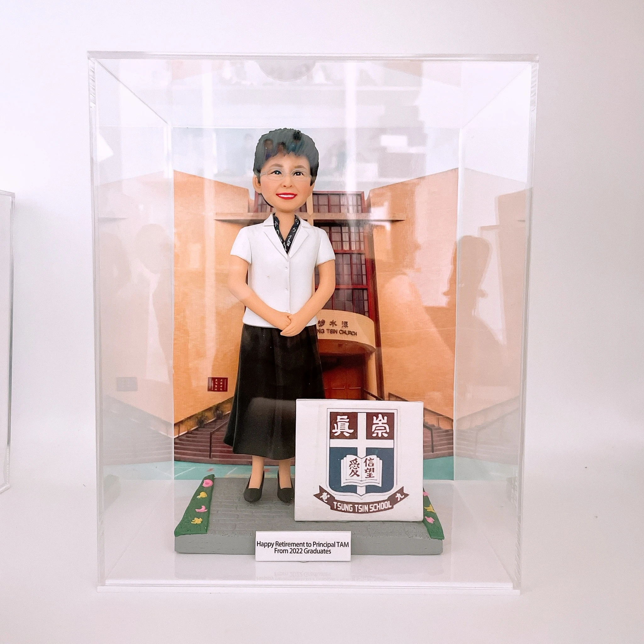 透明壓克力展示盒內，一尊女性人像公仔站在灰色地磚場景中央，穿白色短袖上衣、黑色長裙與黑色平底鞋，短髮、配戴眼鏡並面帶微笑，雙手於身前交握。背板是杏橙色外牆與拱形入口的學校或機構建築。公仔右側有一個立方形立牌，印有校徽與「TSUNG TSIN SCHOOL」字樣；底座前緣的小牌寫着「Happy Retirement to Principal TAM From 2022 Graduates」。地面兩側以綠地與小花點綴。