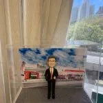戶外企業場景的人像公仔：中年男性穿黑色西裝與紅色領帶，戴眼鏡微笑比讚，站在灰色石板路面底座上；背板為中油站/加油站樣式的大型場景，可見紅白建築、加油島與車道，以及藍天白雲與城市高樓天際線。整體置於透明展示盒內，呈現能源/石油零售產業形象的人像公仔。