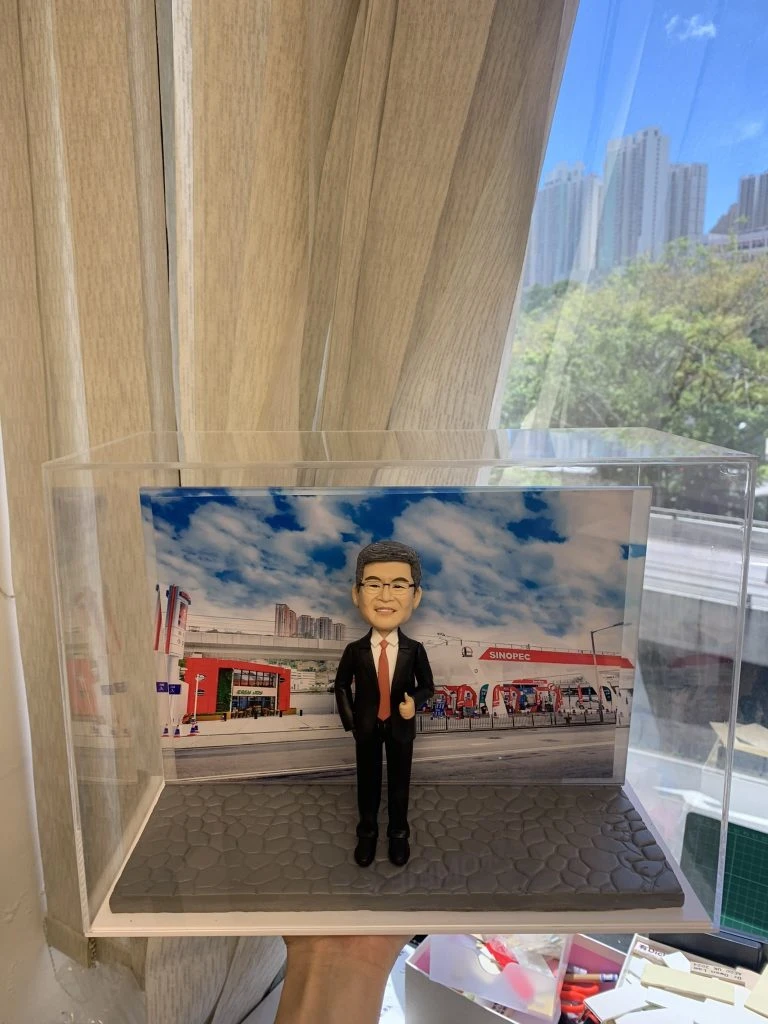 戶外企業場景的人像公仔：中年男性穿黑色西裝與紅色領帶，戴眼鏡微笑比讚，站在灰色石板路面底座上；背板為中油站/加油站樣式的大型場景，可見紅白建築、加油島與車道，以及藍天白雲與城市高樓天際線。整體置於透明展示盒內，呈現能源/石油零售產業形象的人像公仔。