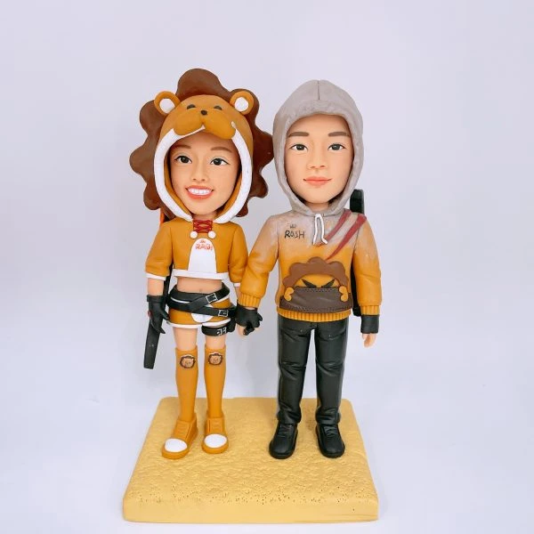 一對情侶主題人像公仔並肩站在淺黃色方形底座上：左側女性穿獅子頭造型連帽與棕色鬃毛、黃橘色短版上衣與短褲，腿上貼有獅子貼布，腳穿長筒襪與球鞋，腰配皮帶與小刀具道具；右側男性著灰色連帽外套，胸前有獅子圖樣，黑色長褲與靴子，背後背帶配件可見。兩人牽手，色調以黃橘與棕、灰為主，背景純白。