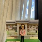 一款老師主題的人像公仔置於校園場景底座：公仔留短髮、戴眼鏡，穿粉色外套、印花上衣與黑色長褲，雙手於身前交握；腳踏灰色地磚，站在綠色草地底座上。背景板為幼稚園/兒童學校門口與窗格，窗內有彩繪與兒童圖案。前方立有中文祝福牌匾，內容祝賀校長退休；右側有小花籃與白色小裝飾物。整體陳列在室內、靠近落地窗與窗簾。