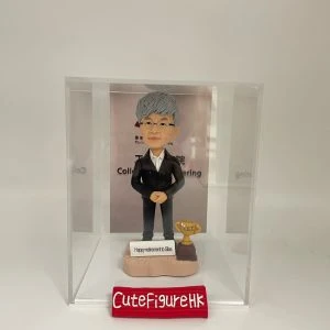 退休老師禮物人像公仔，灰白短髮，戴眼鏡，身穿黑色西裝配白色恤衫，雙手交疊站於淺色底座，底座刻有「Happy retirement to Siles」字樣，旁邊放置金色獎盃，收藏於透明展示盒內。