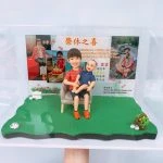 退休主題的人像公仔場景：短髮女性公仔穿紅色格紋上衣與黑裙，腳踩橘色鞋，坐在米色單人椅上懷抱笑臉嬰兒公仔；底座為草地造型，右前方有小花籃與石頭擺飾，左側有白色卷軸。背板為「榮休之喜」主題相片牆，包含多張生活照與感謝字句，呈現同事祝福、回顧服務年資與退休快樂氛圍的人像公仔。