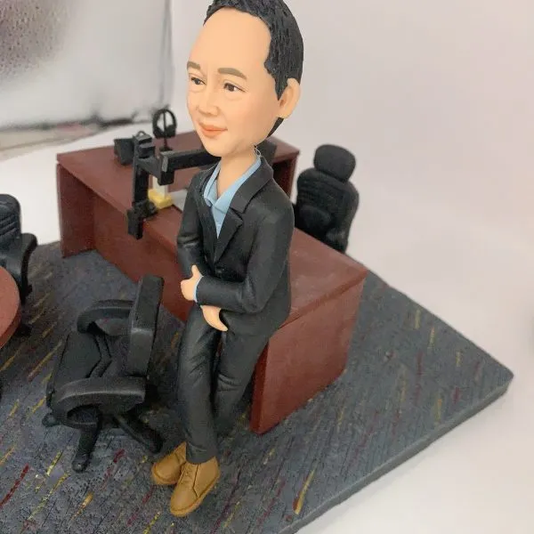 人像公仔辦公室場景：短髮男性公仔穿深色西裝外套與長褲、內搭淺藍襯衫，腳穿棕色皮靴，側身倚靠木質主管桌，雙手自然交疊。桌上配置筆電、螢幕支架、電話座與文具收納；一旁有黑色辦公椅與圓形會議桌，地面為帶有彩色線條的地毯紋理。整體呈現主管/經理級專業辦公氛圍的人像公仔擺設。