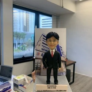 老師禮物人像公仔，人物黑髮，身穿黑色西裝配白色恤衫及紅色領帶，雙手自然放在身側，站立於灰色方形底座，上有金屬銘牌刻有文字，背板印有學校名稱。