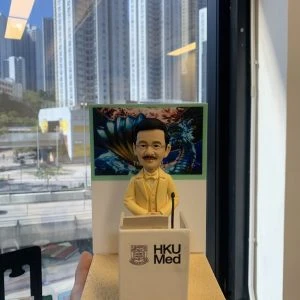 老師禮物人像公仔,人物戴眼鏡、留短鬚,身穿黃色西裝,站在印有「HKU Med」字樣的講台後方,雙手放在講台上,神態莊重。