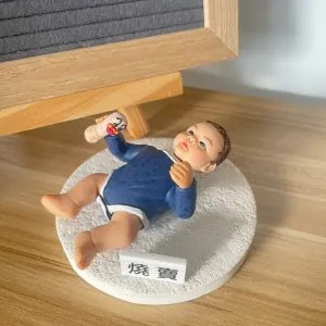 木紋桌面上擺放一款小朋友主題bb人像公仔：嬰兒造型側躺在白色圓形凹凸紋理底座上，穿深藍長袖上衣與白色尿布褲，右手握著奶瓶圖案的小道具，臉頰紅潤、表情放鬆；底座前方有小牌寫著「燒賓」。背景為木框黑灰字母板與牆面。