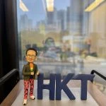 香港電訊(HKT)特色人像公仔，高層人物造型，戴黑框眼鏡，着綠色外套配彩色橙色上衣，紅色長褲印有黑色字樣，腳踏白鞋，站在灰色石紋底座，身旁有深藍色「HKT」立體字母，適合作為高層禮物或公司紀念品。