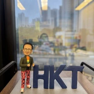 香港電訊(HKT)特色人像公仔，高層人物造型，戴黑框眼鏡，着綠色外套配彩色橙色上衣，紅色長褲印有黑色字樣，腳踏白鞋，站在灰色石紋底座，身旁有深藍色「HKT」立體字母，適合作為高層禮物或公司紀念品。
