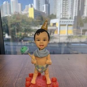 放在木桌上的小朋友主題人像公仔：bb戴著金色尖頂生日帽，上有白色數字1與星星；頸部圍淡藍披巾，下身穿菱格紋尿布褲，雙手各持棒與搖鈴狀道具，赤腳站在紅色愛心形底座上，周圍點綴紅玫瑰造型；背後窗外可見高樓林立與陽光樹影的城市景色。