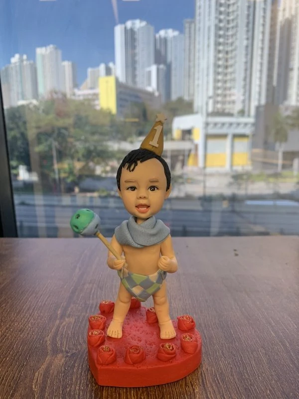 放在木桌上的小朋友主題人像公仔：bb戴著金色尖頂生日帽，上有白色數字1與星星；頸部圍淡藍披巾，下身穿菱格紋尿布褲，雙手各持棒與搖鈴狀道具，赤腳站在紅色愛心形底座上，周圍點綴紅玫瑰造型；背後窗外可見高樓林立與陽光樹影的城市景色。