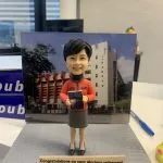 一款歷史老師主題的人像公仔站在灰色紋理底座上，身穿橙色上衣與深灰色半身裙，雙手捧著一本封面寫有「WORLD HISTORY」的書本。背後立有校園建築背景板，可見白色外牆與十字架標誌。底座前方有英文祝賀牌寫著「Congratulations on your glorious retirement Miss Hui」。桌面場景上還有藍色原子筆與辦公用品。