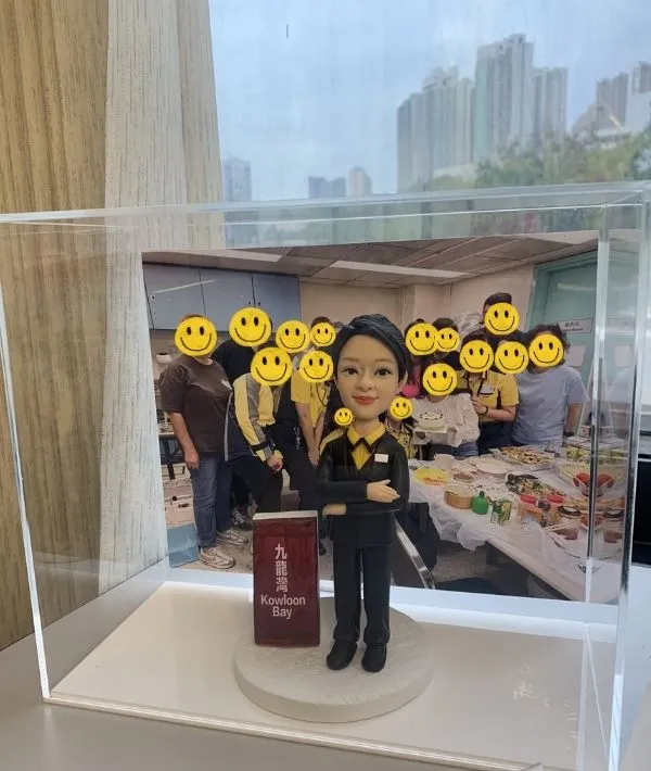 透明壓克力展示盒內，一個穿黑色西裝、黃色襯衫的人像公仔雙手交疊站立，腳下為白色圓形底座；旁邊立著寫有「九龍灣 Kowloon Bay」的紅色站牌造型。展示盒後方放置一張團體合照，十多位穿黃黑制服與便服的人在辦公室/茶水間前合影，桌面擺滿食物與飲品。窗外可見高樓天際線與陰天。