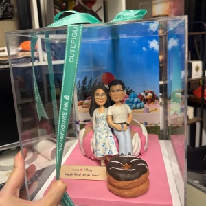 透明禮盒內的一組情侶人像公仔：粉紅方形底座上放置粉白相間的巨型冬甩造型坐墊，情侶並肩坐在上面；左側角色穿白底藍花圖案洋裝、戴眼鏡，右側角色穿白T與淺色長褲。前景有巧克力糖霜巨型冬甩道具，底部小木牌刻有「Joshua ♥ Tiffany Happy birthday! I love you forever!」字樣；背板是糖果甜點主題場景。禮盒以綠色CUTEFIGURE HK緞帶固定，放於室內工作檯面。