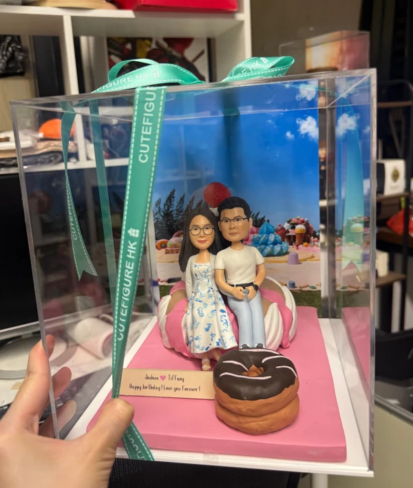 透明禮盒內的一組情侶人像公仔：粉紅方形底座上放置粉白相間的巨型冬甩造型坐墊，情侶並肩坐在上面；左側角色穿白底藍花圖案洋裝、戴眼鏡，右側角色穿白T與淺色長褲。前景有巧克力糖霜巨型冬甩道具，底部小木牌刻有「Joshua ♥ Tiffany Happy birthday! I love you forever!」字樣；背板是糖果甜點主題場景。禮盒以綠色CUTEFIGURE HK緞帶固定，放於室內工作檯面。