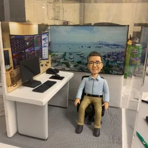 坐在辦公椅上的人像公仔場景：中年男性造型，戴黑框眼鏡，穿淺藍襯衫與卡其長褲、深色鞋。前方白色L形辦公桌擺有電腦螢幕、鍵盤、滑鼠與數據單；左側背板為多屏交易數據與K線走勢圖，右側背板為臨海城市港口景觀。整體置於透明展示盒內，地面為灰色顆粒質感。呈現金融交易/投資分析工作氛圍的人像公仔。