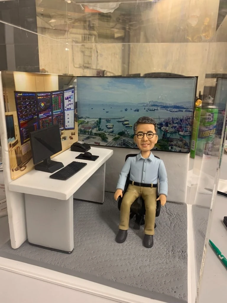 坐在辦公椅上的人像公仔場景：中年男性造型，戴黑框眼鏡，穿淺藍襯衫與卡其長褲、深色鞋。前方白色L形辦公桌擺有電腦螢幕、鍵盤、滑鼠與數據單；左側背板為多屏交易數據與K線走勢圖，右側背板為臨海城市港口景觀。整體置於透明展示盒內，地面為灰色顆粒質感。呈現金融交易/投資分析工作氛圍的人像公仔。