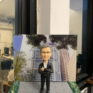 高層榮休主題的人像公仔：灰髮戴眼鏡的男士身穿黑色西裝與白襯衫，雙手抱胸自信站立於灰色磚紋地台中央；腳邊左前方立有白色名牌，上印「Jimmy Gor｜Happy Retirement」字樣。背板為都會金融區高樓與樹蔭交錯的城市景觀，象徵商務領域的職涯背景；地台兩側加入綠地灑花紋理，增添儀式感。整體以室內靠窗拍攝，光線清朗，呈現專業、尊榮且具紀念價值的企業高管人像公仔。