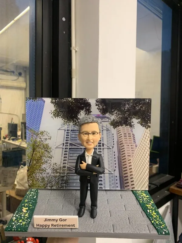高層榮休主題的人像公仔：灰髮戴眼鏡的男士身穿黑色西裝與白襯衫，雙手抱胸自信站立於灰色磚紋地台中央；腳邊左前方立有白色名牌，上印「Jimmy Gor｜Happy Retirement」字樣。背板為都會金融區高樓與樹蔭交錯的城市景觀，象徵商務領域的職涯背景；地台兩側加入綠地灑花紋理，增添儀式感。整體以室內靠窗拍攝，光線清朗，呈現專業、尊榮且具紀念價值的企業高管人像公仔。