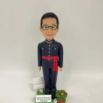 身穿深藍色制服的人像公仔直立於草地質感底座上；制服為立領、肩章與口袋翻蓋設計，胸前有金屬鏈與扣件，褲側有紅色側條，腰間束紅色流蘇腰帶，腳穿黑皮鞋；公仔短髮、戴黑框眼鏡、微笑。底座左後有白色告示牌與銀色配件，右前有編織籃盛放綠葉與黃色花；前置銘牌寫著 “Happy Retirement Mr Francis FONG May 2022”。