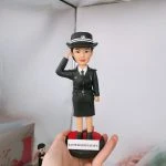 一款女性懲教風格的人像公仔站在黑紅圓形底座上，右手抬至帽沿敬禮；身穿深色制服外套配裙裝、白色襯衫與深色領帶，胸前有名牌與勛章，肩章與腰帶細節清晰；頭戴深色硬帽，帽緣有白色橫紋與徽章；底座前貼有白色標籤文字。背景為室內角落，一隻手掌托住底座。