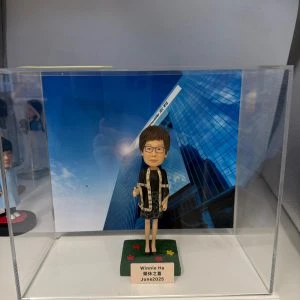 女上司形象的人像公仔放置於透明展示盒內，背景為高樓大廈與藍天照片；公仔戴眼鏡、短髮，身穿黑色連身裙與黑色外套，外套邊緣有淺色滾邊與花紋細節，腳穿黑鞋，右手比讚姿勢。公仔站在綠色草地質感的方形底座上，底座灑有紅黃花形裝飾；前方小銘牌寫有「Winnie Ha 榮休之喜 June 2025」。展示盒左側可見另一公仔的局部。