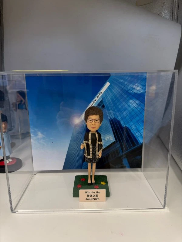 女上司形象的人像公仔放置於透明展示盒內，背景為高樓大廈與藍天照片；公仔戴眼鏡、短髮，身穿黑色連身裙與黑色外套，外套邊緣有淺色滾邊與花紋細節，腳穿黑鞋，右手比讚姿勢。公仔站在綠色草地質感的方形底座上，底座灑有紅黃花形裝飾；前方小銘牌寫有「Winnie Ha 榮休之喜 June 2025」。展示盒左側可見另一公仔的局部。