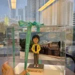 透明展示盒內的人像公仔站立於方形底座上：角色穿黃色西裝外套、黑色上衣與灰色長褲，雙手交疊於身前；身後放置學校外牆與校門的相片式背景。展示盒以綠色CUTEFIGURE HK緞帶綁結，前方名牌印有中英文職銜與名稱字樣；盒子置於木桌上，窗外雨滴與高樓景象映入玻璃。