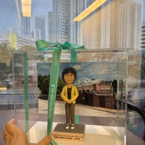 透明展示盒內的人像公仔站立於方形底座上：角色穿黃色西裝外套、黑色上衣與灰色長褲，雙手交疊於身前；身後放置學校外牆與校門的相片式背景。展示盒以綠色CUTEFIGURE HK緞帶綁結，前方名牌印有中英文職銜與名稱字樣；盒子置於木桌上，窗外雨滴與高樓景象映入玻璃。