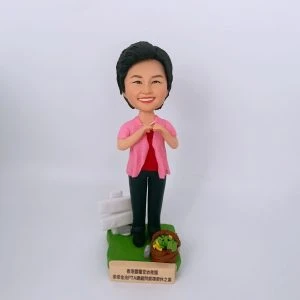 女士主題的人像公仔站在綠色草地質感的長方形底座上，短髮微笑，穿粉紅色開襟外套搭配紅色上衣、深色長褲與黑鞋；雙手於胸前比出愛心手勢。底座左後方有白色告示牌裝飾，右前方有裝著綠色蔬菜的小籃子；前置金色銘牌刻有「香港聖道堂幼稚園 路德金池PTA顧問榮休誌喜」等字樣。