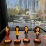 四款教師人像公仔排成一列放在木面桌上，背後是落地窗與高樓城市景。每個公仔立於黑紅圓形底座並配白色名牌：左起角色穿酒紅套裝；第二位穿黃色外套與深色褲；第三位戴眼鏡、深色外套搭配藍白格紋裙，手持小卡片；第四位長髮、穿灰色上衣配紅心別針並持卡片。名牌上以英中標示職銜與年份。整體呈現學校團隊展示氛圍。