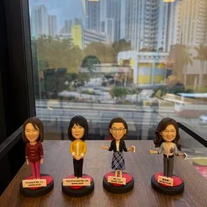 四款教師人像公仔排成一列放在木面桌上，背後是落地窗與高樓城市景。每個公仔立於黑紅圓形底座並配白色名牌：左起角色穿酒紅套裝；第二位穿黃色外套與深色褲；第三位戴眼鏡、深色外套搭配藍白格紋裙，手持小卡片；第四位長髮、穿灰色上衣配紅心別針並持卡片。名牌上以英中標示職銜與年份。整體呈現學校團隊展示氛圍。