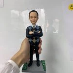 男性人像公仔站在灰色石磚紋方形底座上，底座兩側綠色草地帶白色小花點綴；公仔雙臂交叉於胸前，面露微笑，穿藍黑色菱格（Argyle）針織背心、淺藍襯衫與酒紅色領帶，下著深色長褲與黑皮鞋，左手腕配戴手錶。背景為室內白色牆面與反光，一隻手掌托住底座。