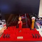 一座商業主題的人像公仔場景：中央是著紅色長禮服、手持麥克風的女性公仔，站在紅色絨面舞台上。舞台前景以立體字樣拼出「INV Sales Team」，兩側各有三支黑色話筒支架。右後方立有帶紅白幾何標誌的圓形獎杯柱，底座寫有「HSBC」。後方背景板呈現夜色城市與霓虹燈廣告。前方名牌寫著「Winnie Ng」。整體置於工作檯面上拍攝。