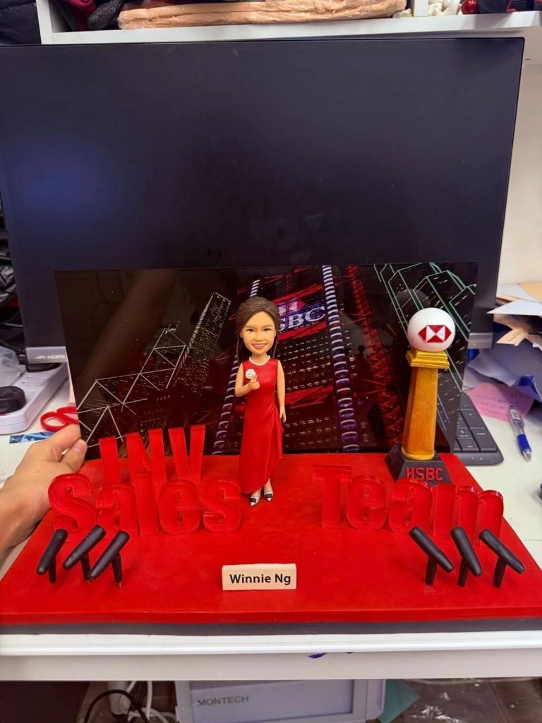 一座商業主題的人像公仔場景：中央是著紅色長禮服、手持麥克風的女性公仔，站在紅色絨面舞台上。舞台前景以立體字樣拼出「INV Sales Team」，兩側各有三支黑色話筒支架。右後方立有帶紅白幾何標誌的圓形獎杯柱，底座寫有「HSBC」。後方背景板呈現夜色城市與霓虹燈廣告。前方名牌寫著「Winnie Ng」。整體置於工作檯面上拍攝。