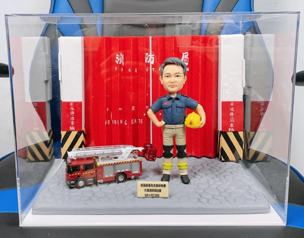 透明展示盒內的消防主題人像公仔場景：人物穿深藍上衣與卡其色防護褲、黑色護膝與靴子，右手叉腰、左手握黃色消防頭盔，站在灰色石板紋理台座上。背景為紅色摺疊式消防局大門，兩側有黑黃斜紋防撞柱與白色牆面，上有「消防局 FIRE STATION」字樣。人物左前方停放一輛紅色雲梯消防車模型。台座前方置有金色小銘牌，刻有祝福與代號文字。整組作品置於透明壓克力展示盒中，外部可見藍黑色椅背。
