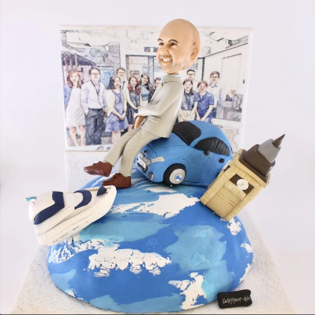 桌面尺寸手作擺設：藍白相間的地球基座上，右側傾斜插入大笨鐘造型的立體建築小模型；左前側放著白藍配色的小型郵輪；中央是一位穿淺灰西裝、棕色皮鞋的人像公仔，微笑坐在一台藍色小車上，身後以團隊合照風格的插畫作為背景。整體象徵從英國與海上旅程出發，作為退休禮物或移民禮物的祝福。