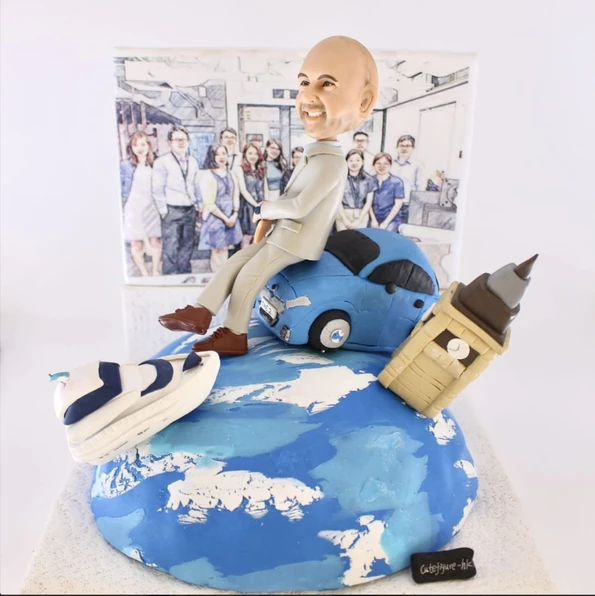 桌面尺寸手作擺設：藍白相間的地球基座上，右側傾斜插入大笨鐘造型的立體建築小模型；左前側放著白藍配色的小型郵輪；中央是一位穿淺灰西裝、棕色皮鞋的人像公仔，微笑坐在一台藍色小車上，身後以團隊合照風格的插畫作為背景。整體象徵從英國與海上旅程出發，作為退休禮物或移民禮物的祝福。