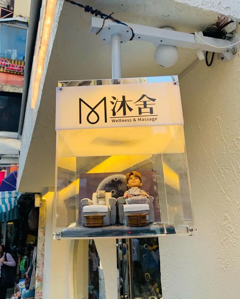多張照片展示同一套開張人像公仔：一位穿著淺灰藍浴袍的Q版人物坐在按摩足浴沙發上，腳前有絨面腳墊與兩個木質足浴桶，旁邊另置一張同款空沙發與托盤小茶杯。背景板為灰色圓形圖案，整體置於灰白色地台。作品被放入透明燈箱中，燈箱上印有「沐舍 Wellness & Massage」字樣，固定在店舖外牆作為開張或門面擺設；遠景可見店鋪招牌、圓形窗與街景。