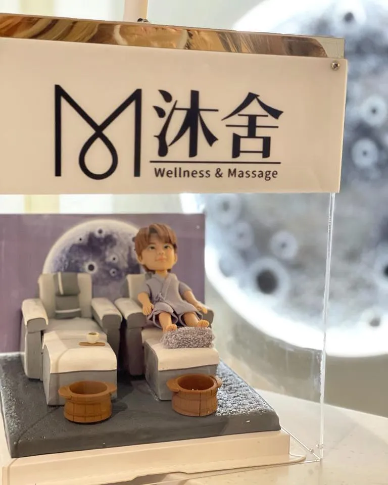 多張照片展示同一套開張人像公仔：一位穿著淺灰藍浴袍的Q版人物坐在按摩足浴沙發上，腳前有絨面腳墊與兩個木質足浴桶，旁邊另置一張同款空沙發與托盤小茶杯。背景板為灰色圓形圖案，整體置於灰白色地台。作品被放入透明燈箱中，燈箱上印有「沐舍 Wellness & Massage」字樣，固定在店舖外牆作為開張或門面擺設；遠景可見店鋪招牌、圓形窗與街景。