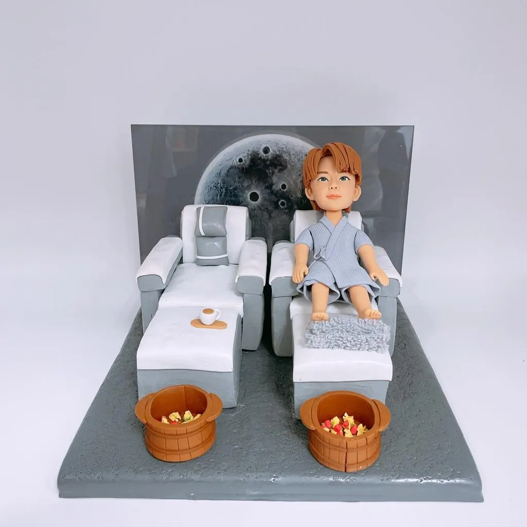 多張照片展示同一套開張人像公仔：一位穿著淺灰藍浴袍的Q版人物坐在按摩足浴沙發上，腳前有絨面腳墊與兩個木質足浴桶，旁邊另置一張同款空沙發與托盤小茶杯。背景板為灰色圓形圖案，整體置於灰白色地台。作品被放入透明燈箱中，燈箱上印有「沐舍 Wellness & Massage」字樣，固定在店舖外牆作為開張或門面擺設；遠景可見店鋪招牌、圓形窗與街景。