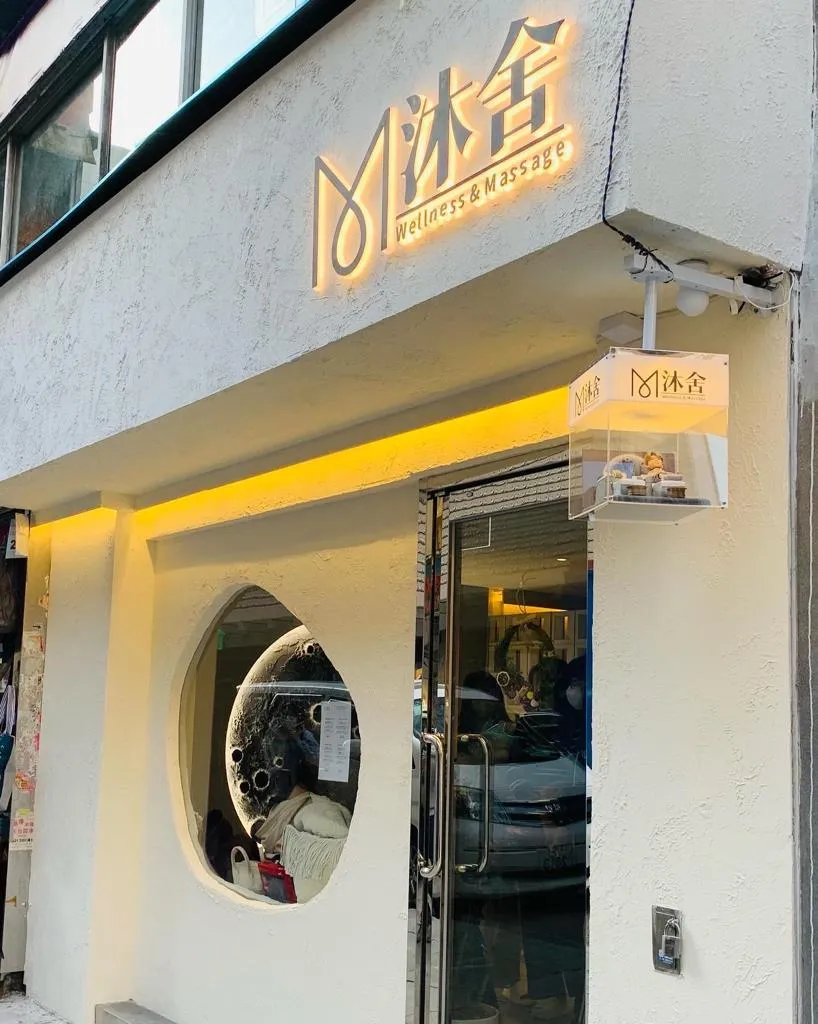 多張照片展示同一套開張人像公仔：一位穿著淺灰藍浴袍的Q版人物坐在按摩足浴沙發上，腳前有絨面腳墊與兩個木質足浴桶，旁邊另置一張同款空沙發與托盤小茶杯。背景板為灰色圓形圖案，整體置於灰白色地台。作品被放入透明燈箱中，燈箱上印有「沐舍 Wellness & Massage」字樣，固定在店舖外牆作為開張或門面擺設；遠景可見店鋪招牌、圓形窗與街景。