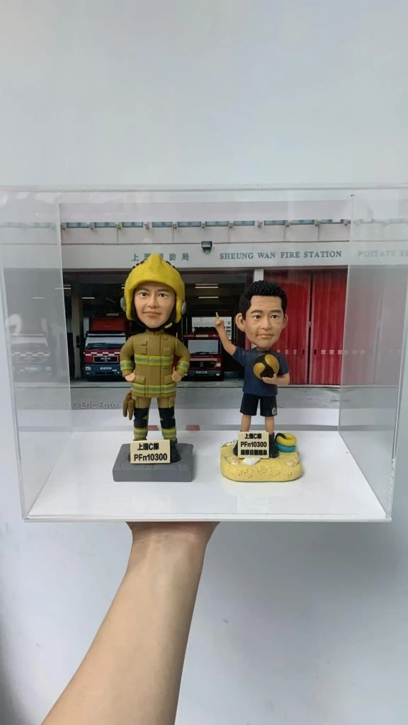 一只透明展示盒被手掌托起，盒內放置兩尊人像公仔，背板為「SHEUNG WAN FIRE STATION／上環消防局」外觀與紅色車庫門。左側公仔穿卡其色消防防護衣與黑黃相間護膝、黑靴，頭戴黃色消防頭盔，雙手叉腰站在灰色方形台座上；台座前方貼有黑字標籤。右側公仔穿深藍上衣與黑色短褲、白鞋，右手食指上指做比劃手勢，左手抱著球狀物，腳邊有黃色與藍色救援圈造型飾件，站在黃色沙地質感台座上；前方同樣有黑字標籤。盒內可見消防車停放的車庫影像細節。