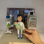 迷你場景中的醫生人像公仔站在灰色地台中央：穿淺藍手術服與白袍，雙手抱著裹綠毯的新生兒，右手持綠色復甦球。左後方是帶螢幕的生命監測儀與移動推車，右側為透明保溫箱模型；背景板呈現病房環境。前緣有淺膚色名牌，印有科室、姓名、職稱與年份字樣，呼應退休紀念主題。
