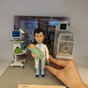 迷你場景中的醫生人像公仔站在灰色地台中央：穿淺藍手術服與白袍，雙手抱著裹綠毯的新生兒，右手持綠色復甦球。左後方是帶螢幕的生命監測儀與移動推車，右側為透明保溫箱模型；背景板呈現病房環境。前緣有淺膚色名牌，印有科室、姓名、職稱與年份字樣，呼應退休紀念主題。