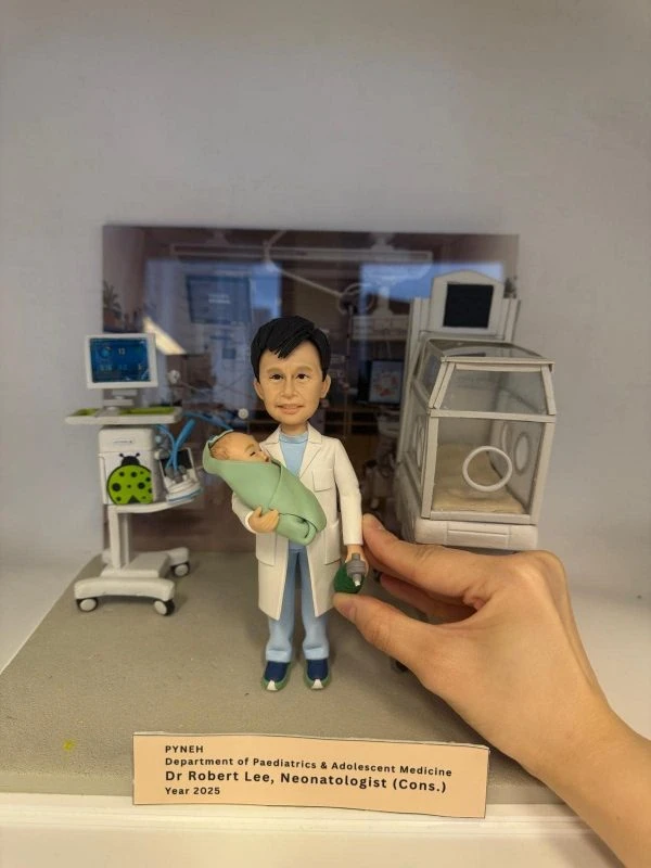 迷你場景中的醫生人像公仔站在灰色地台中央：穿淺藍手術服與白袍，雙手抱著裹綠毯的新生兒，右手持綠色復甦球。左後方是帶螢幕的生命監測儀與移動推車，右側為透明保溫箱模型；背景板呈現病房環境。前緣有淺膚色名牌，印有科室、姓名、職稱與年份字樣，呼應退休紀念主題。