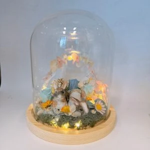 一座玻璃罩擺飾置於淺色木質圓形底座上，罩內鋪有苔蘚與碎石，中央有兩隻兔子動物公仔相依而坐；周圍點綴多朵永生花與小雛菊、藍色與粉色玫瑰，後方以粉橘色系花瓣組成半圓拱門。底座四周有暖黃小燈串發光，整體呈溫暖柔和的夜燈效果。
